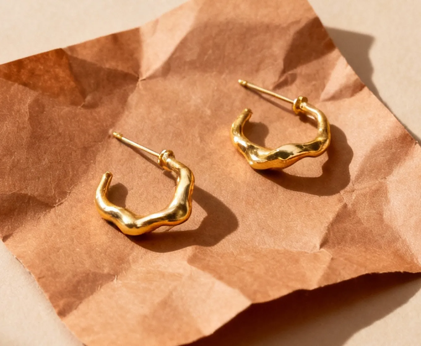 Pair of textured gold hoop earrings with an irregular, wavy shape displayed on crumpled brown paper.