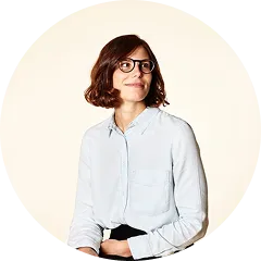 Woman with short brown hair and glasses wearing a light blue button-up shirt against a white background.
